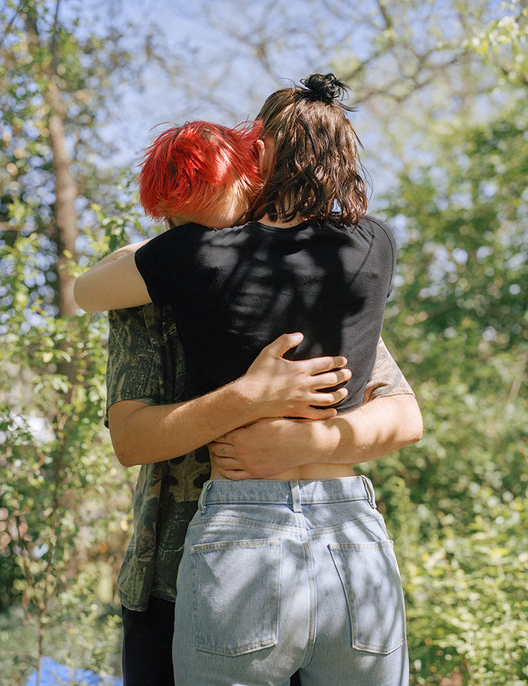 Infinite Tenderness by Peyton Fulford — Two figures embrace outdoors against a backdrop of leafy trees and bright sky.