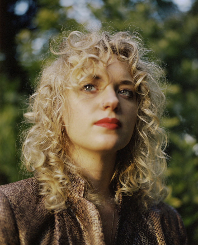 Infinite Tenderness by Peyton Fulford — A portrait photograph featuring a single figure with voluminous blonde curled hair, positioned against a softly blurred green foliage background.