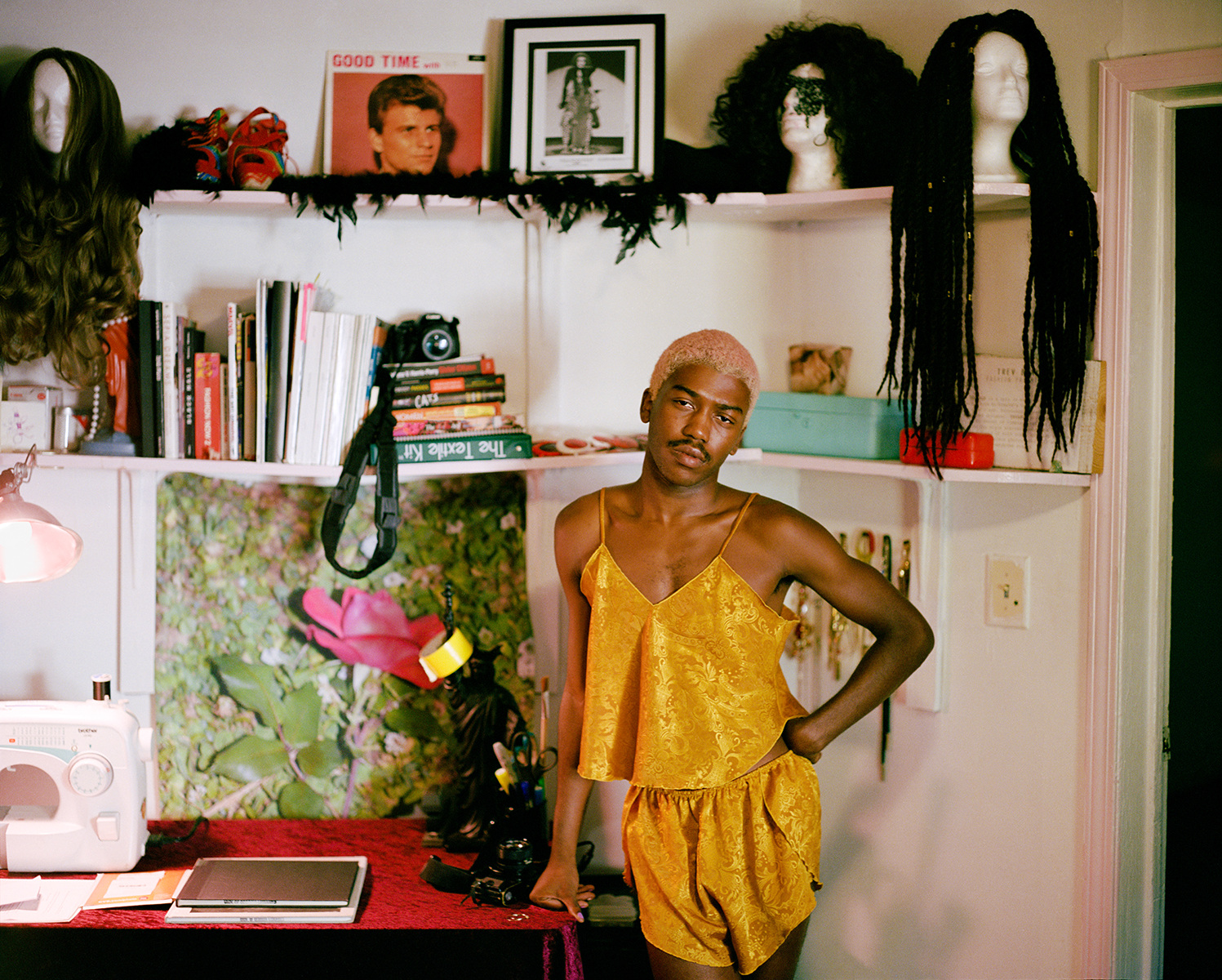 Infinite Tenderness by Peyton Fulford — A figure in a mustard-yellow velvet camisole and matching shorts stands centered against a white shelving unit, one hand on hip in a direct frontal pose.