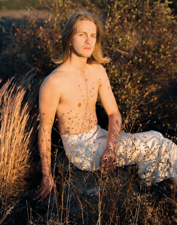 Infinite Tenderness by Peyton Fulford — A shirtless figure with shoulder-length brown hair sits among dried grasses and vegetation in golden-hour light.