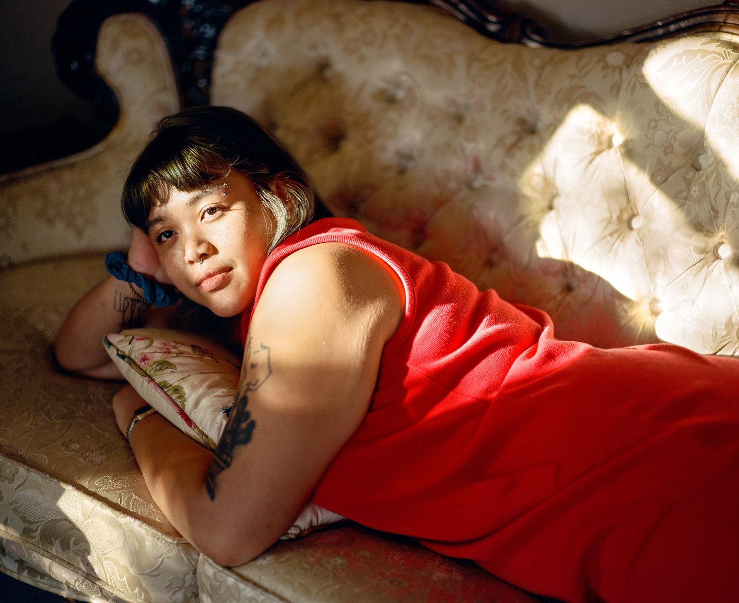 Infinite Tenderness by Peyton Fulford — A figure in a bright red sleeveless shirt lies prone on patterned upholstered furniture, positioned diagonally across the frame.