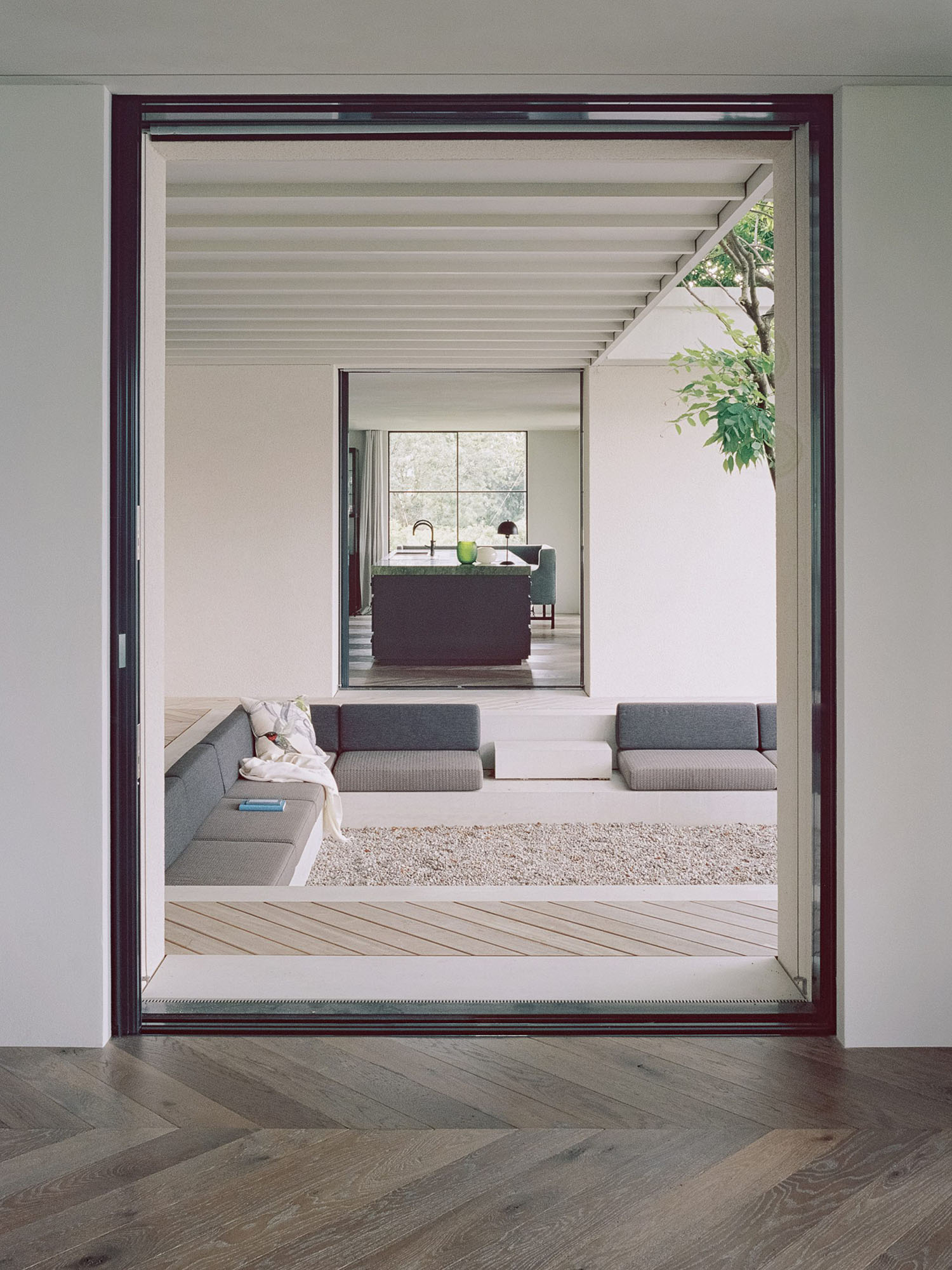 House at a Vineyard by Think Architecture — Viewed from the interior hallway, a threshold framed by dark timber and aluminum reveals a sunken courtyard with pale timber decking and gravel surface.