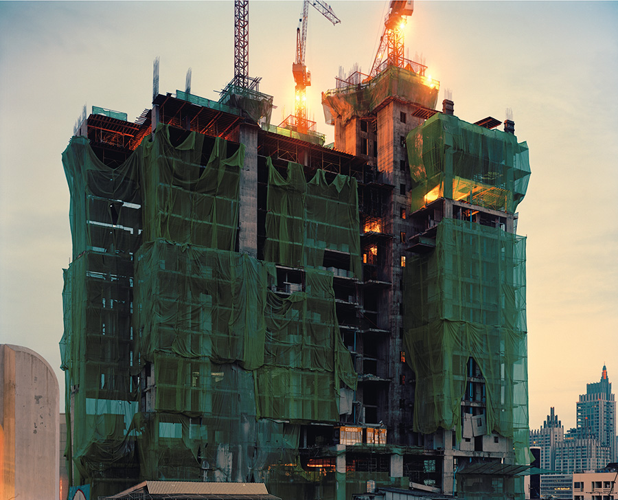 Aftermath by Jörn Vanhöfen — A multi-story building under construction is wrapped entirely in sage-green protective netting, creating a monolithic facade that obscures the structural...