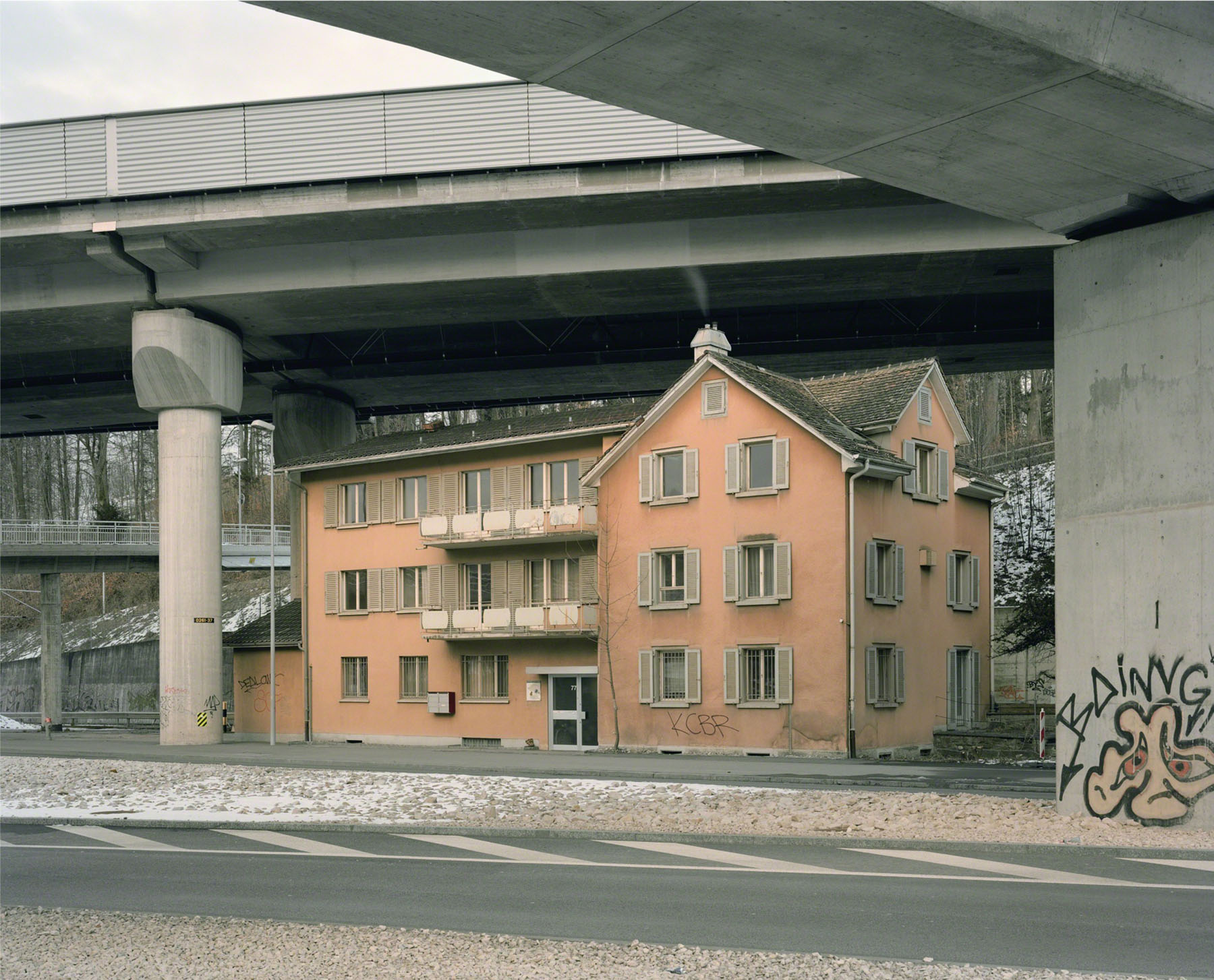 Aftermath by Jörn Vanhöfen — A residential apartment building rendered in terracotta stucco sits beneath a massive concrete highway overpass, its three-story facade dominated by...