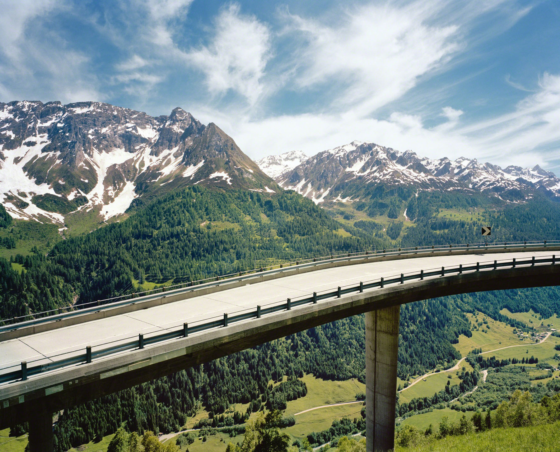 Aftermath by Jörn Vanhöfen — A elevated highway viaduct bisects the alpine valley in a stark horizontal gesture, its concrete deck and steel railings cutting through dense coniferous...