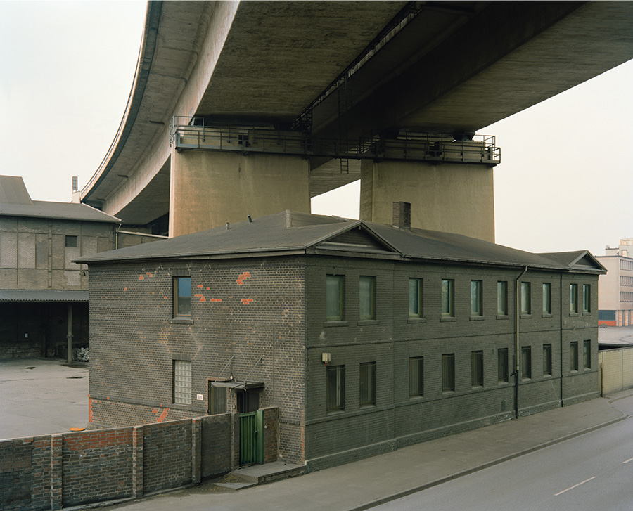Aftermath by Jörn Vanhöfen — A brick industrial building with green-painted window frames and rendered walls sits dwarfed beneath a massive concrete highway overpass.