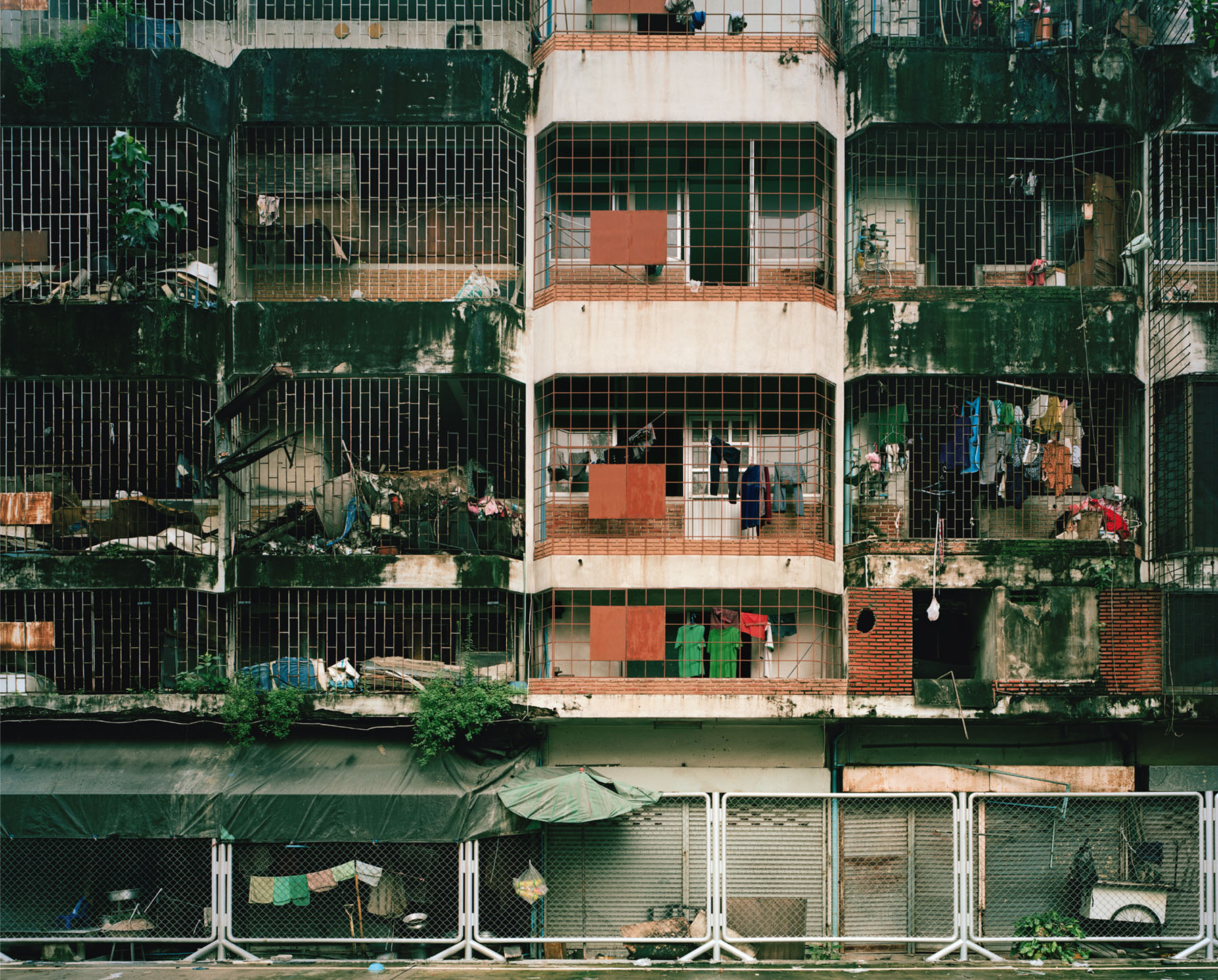 Aftermath by Jörn Vanhöfen — A densely layered residential facade fronts a narrow waterway, its weathered brick and tile surfaces stained with algae and mineral deposits.