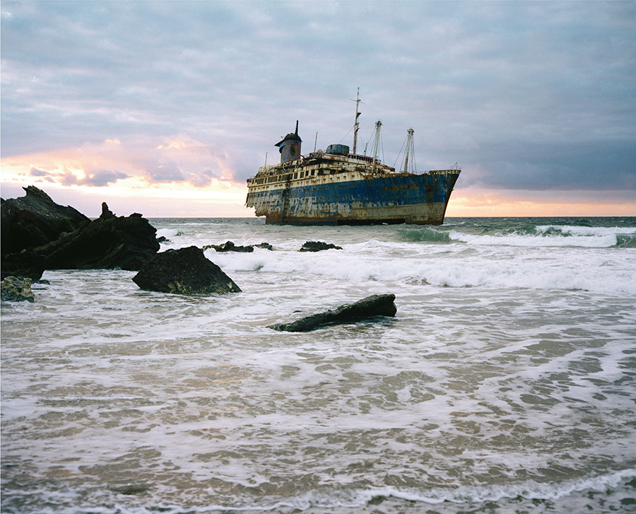 Aftermath by Jörn Vanhöfen — A rusted cargo vessel lists heavily in shallow coastal waters, its blue and yellow hull corroded and deteriorating.