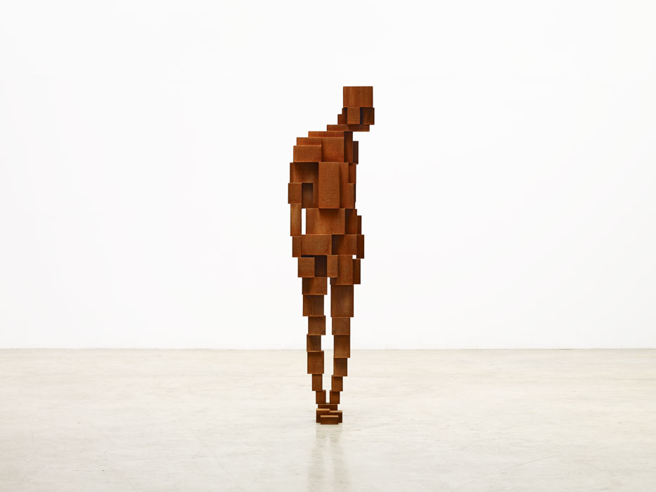 Sculptures by Antony Gormley — A timber-block figure stands in frontal orientation on a polished concrete floor against a white gallery wall.