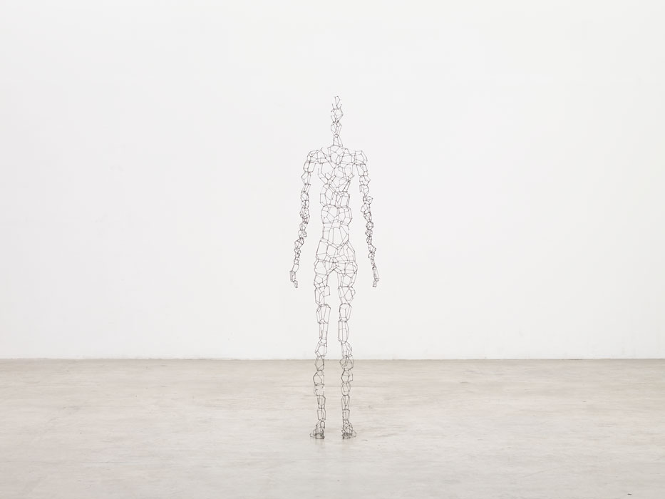 Sculptures by Antony Gormley — A towering wireframe figure constructed from interconnected rectangular timber blocks stands vertically in an expansive concrete gallery space.