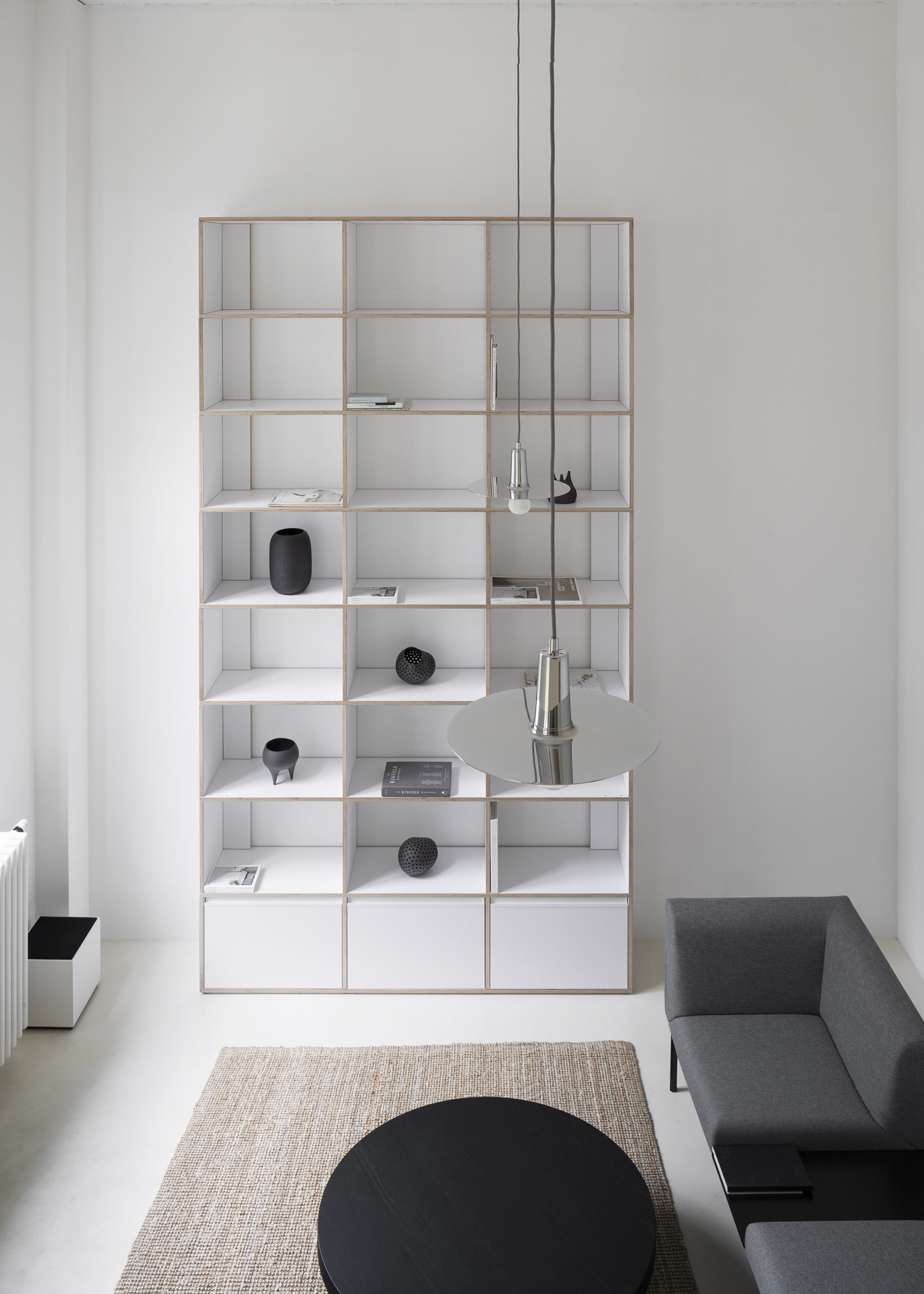 A-PLACE K907 by Thisispaper Studio — A floor-to-ceiling modular shelving unit dominates the residential interior, its timber frame and white plywood compartments creating a rhythmic grid across...