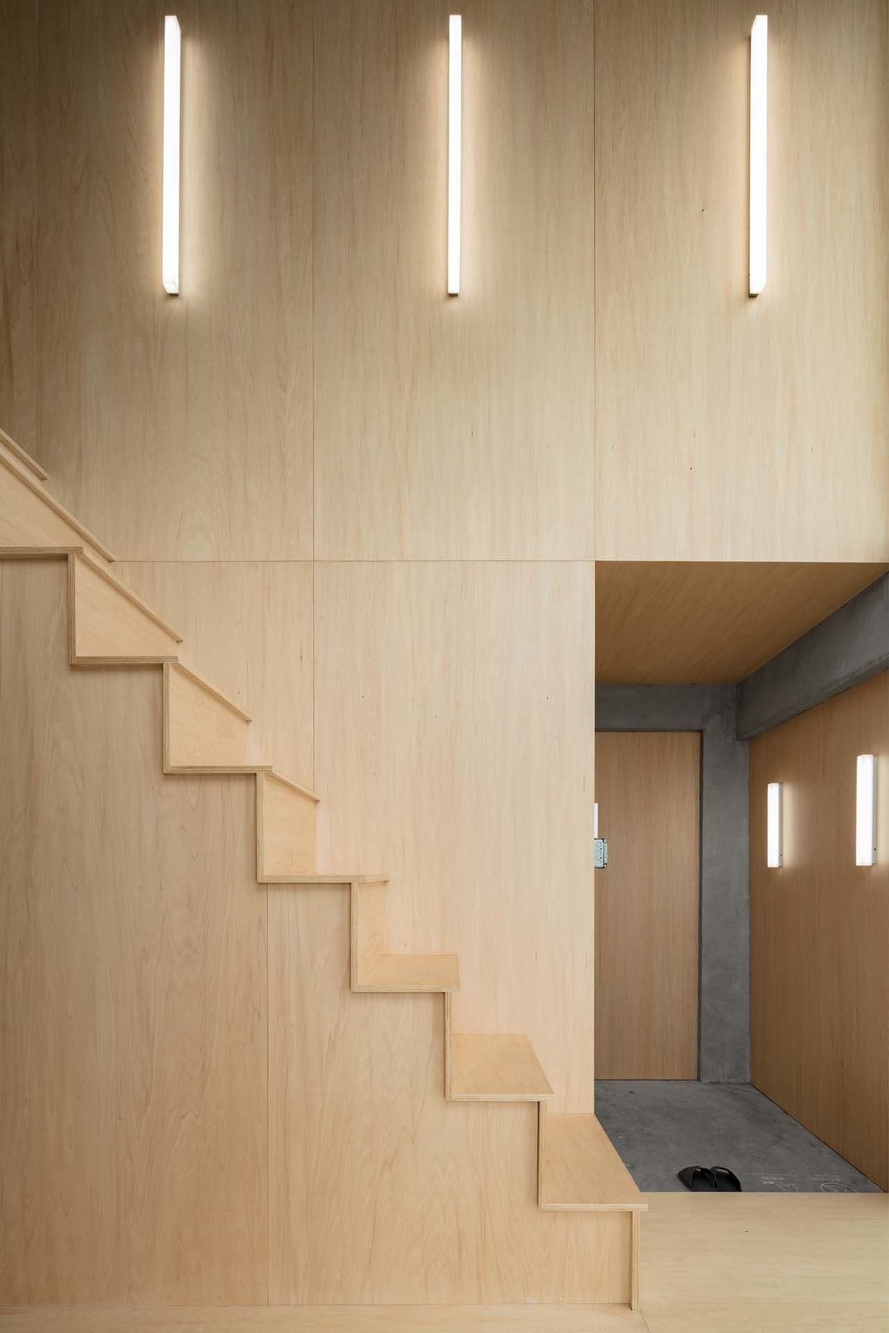 Hotel Far & Near Nanhao St. by kooo architects — A timber-lined staircase ascends the left wall of this double-height interior, its stepped geometry casting incremental shadows across light wood surfaces.