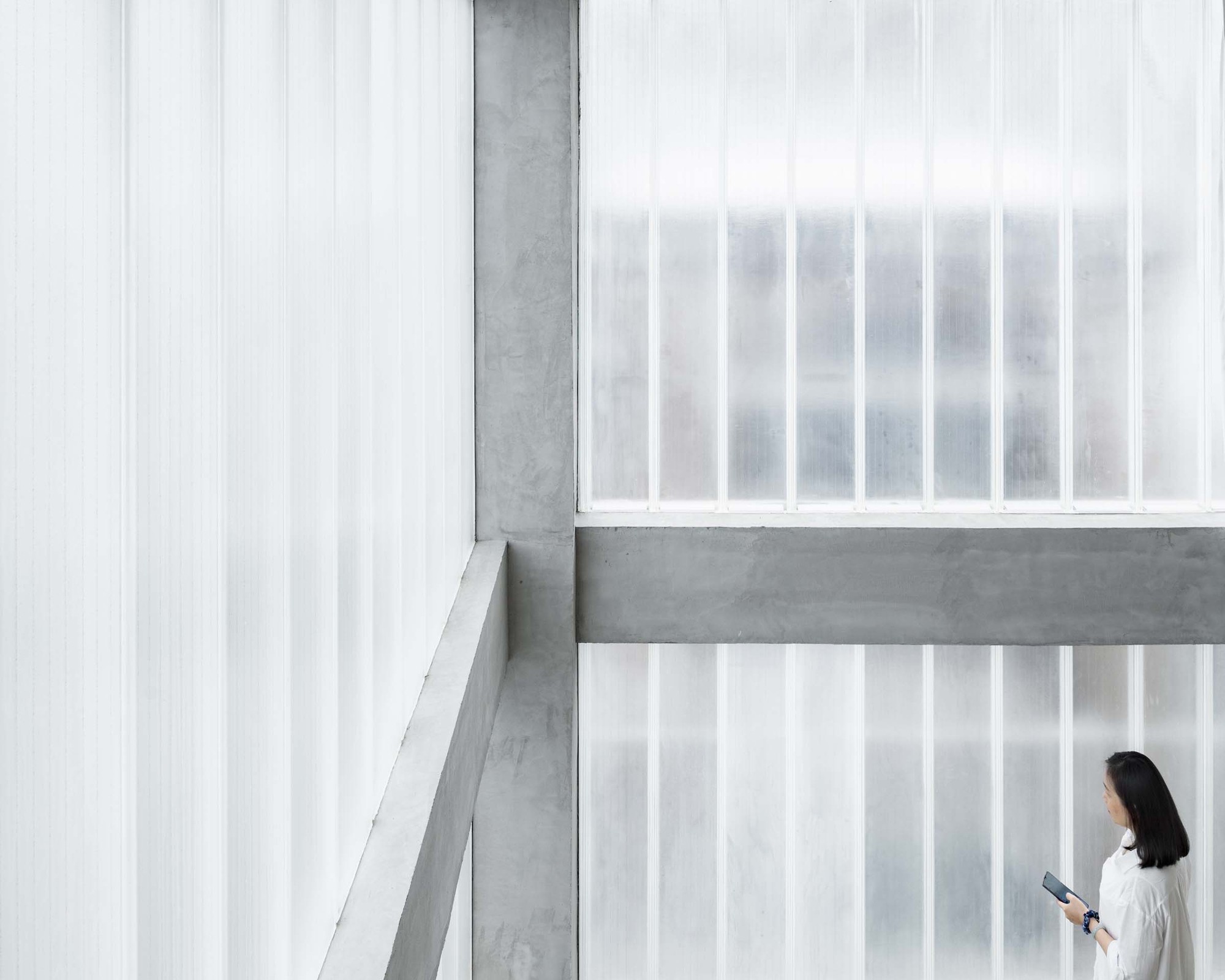 Hotel Far & Near Nanhao St. by kooo architects — A figure in white linen stands in profile within a concrete-framed interior, holding a tablet device.