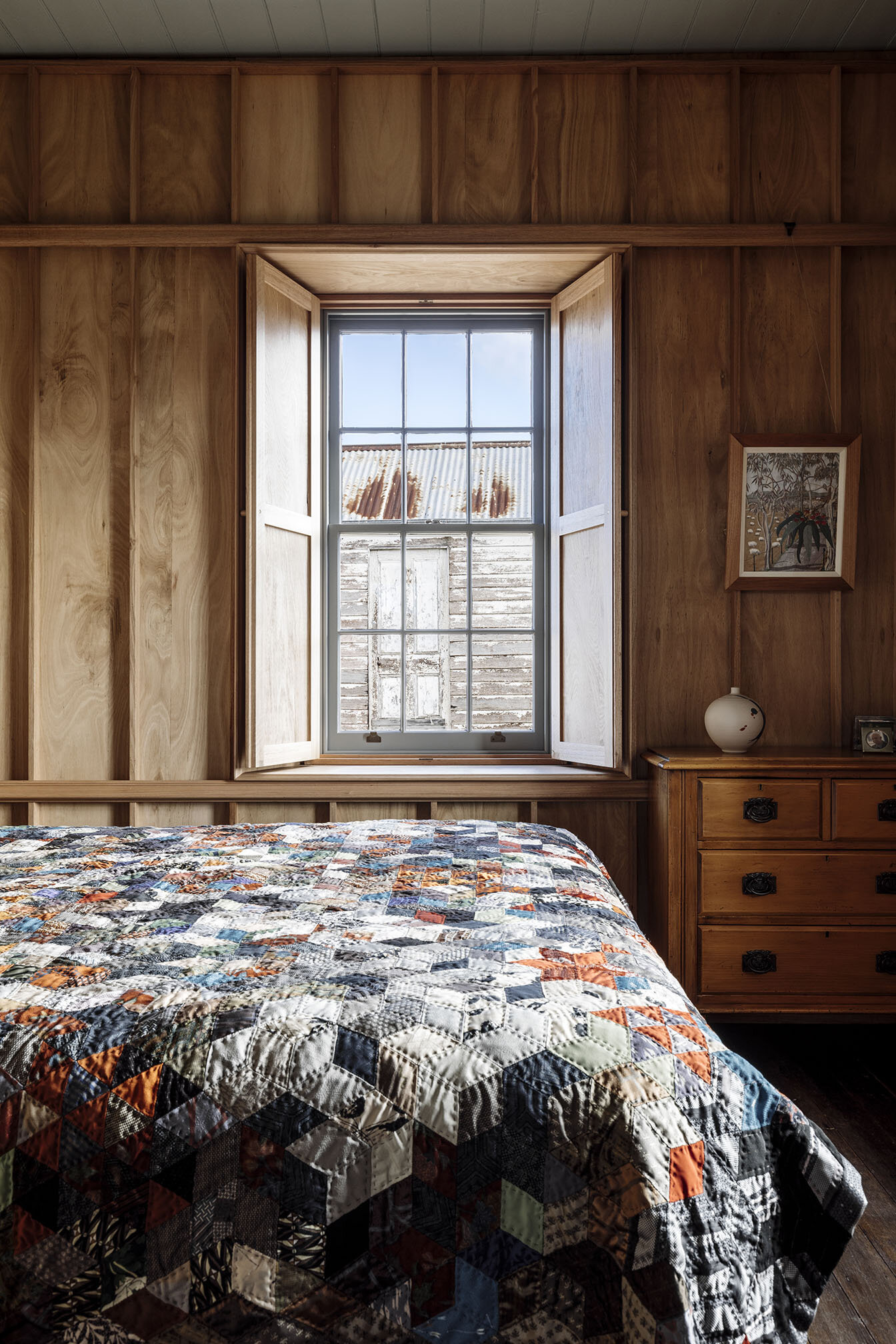 Bozen's Cottage by Taylor + Hinds Architects — A timber-lined bedroom features a recessed window opening with painted white timber reveals, creating spatial depth within richly stained wood wall paneling...