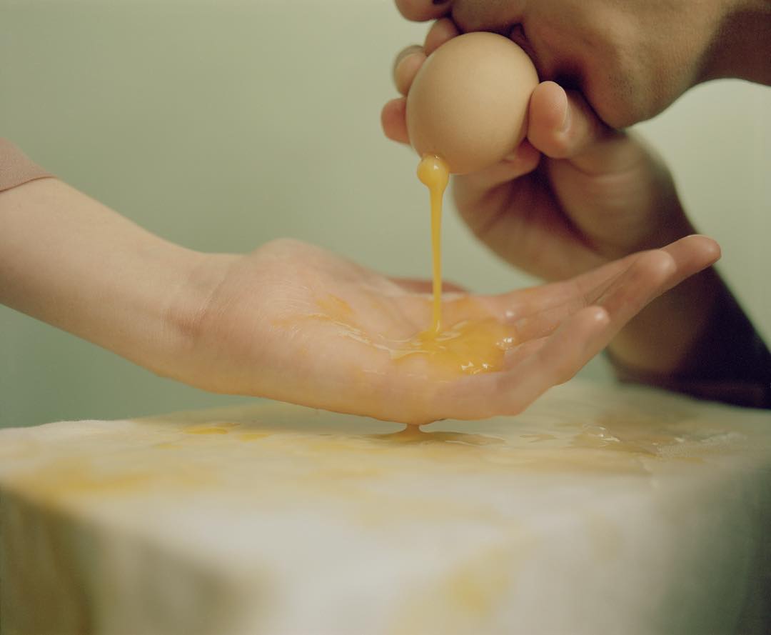 Selected works by András Ladocsi — A child's hand catches raw egg yolk dripping from a cracked shell held above, captured in close-up against a neutral pale green background.