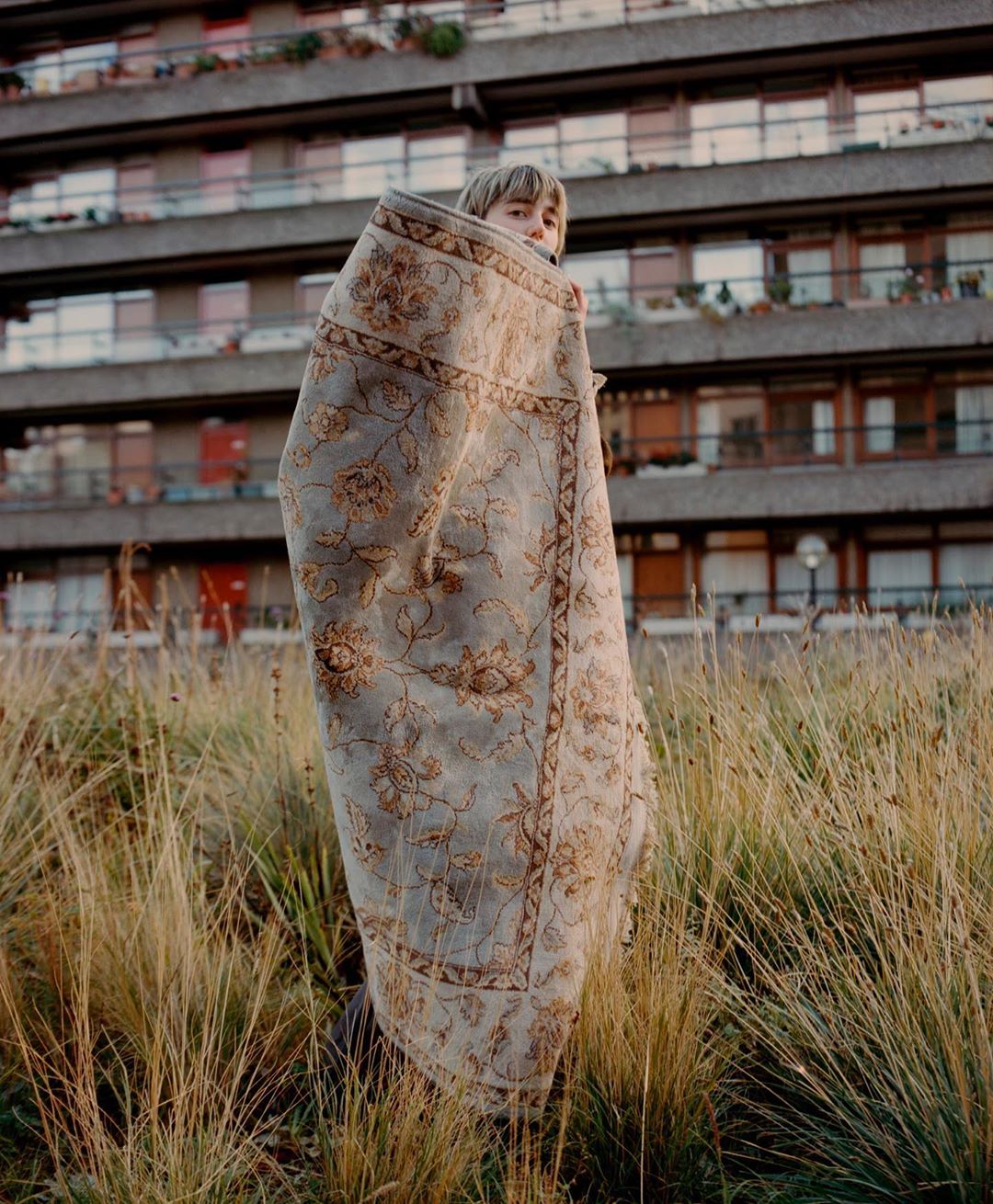 Selected works by András Ladocsi — A child wrapped in a patterned textile stands in dry grass facing the camera, with a modernist apartment building occupying the background.