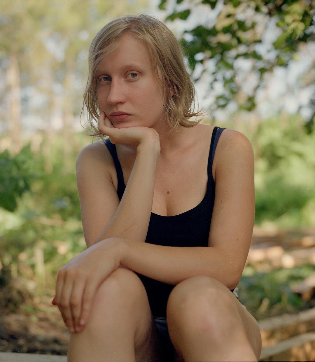 Selected works by András Ladocsi — Portrait of a young woman with shoulder-length blonde hair, positioned in a crouched pose against a blurred garden background with flowering vines and green...