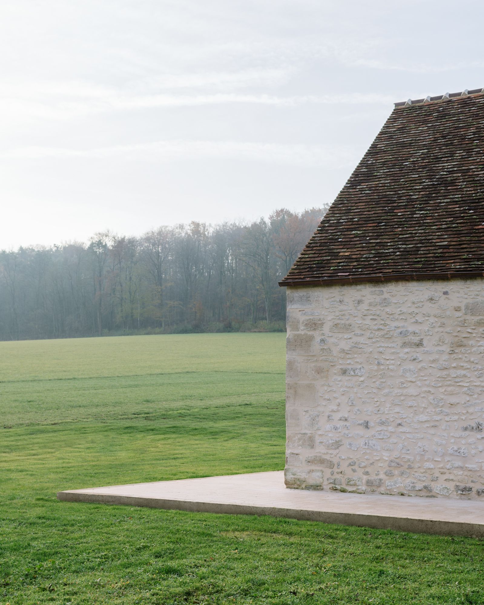 Réhabilitation de Ferme by Collet & Muller Architects — A coursed stone masonry wall with weathered surface patina meets a flush concrete platform that creates a sharp horizontal datum against the sloped terrain.