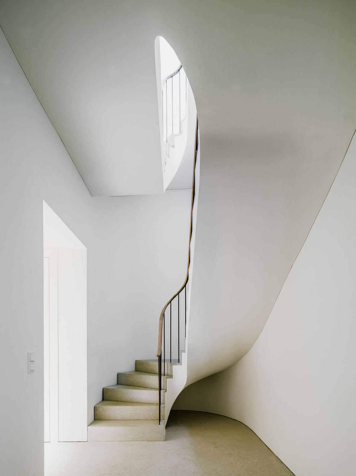 Bogenhausen House by David Chipperfield Architects — A sculptural staircase ascends through a double-height void, its curved white plaster walls creating a continuous spiral form that rises toward a skylight...