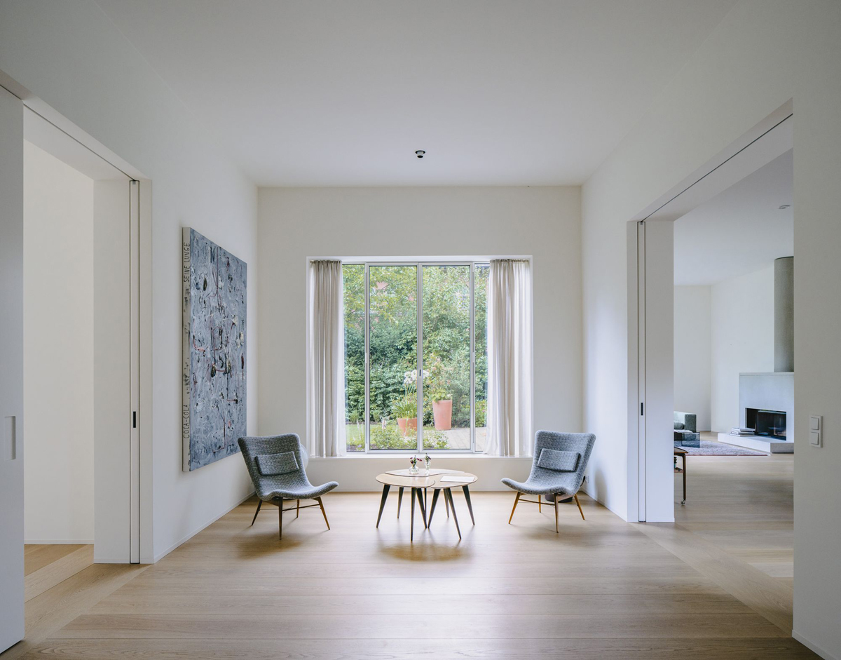 Bogenhausen House by David Chipperfield Architects — A double-height gallery space anchors the Bogenhausen House interior, featuring a 4x2 gridded window with dark aluminum mullions framing views of mature...