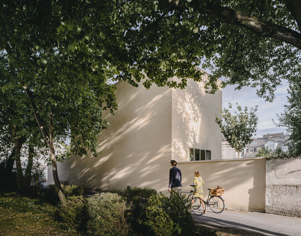 Bogenhausen House by David Chipperfield Architects — Two figures with a bicycle pause at the rendered perimeter wall of Bogenhausen House, framed beneath mature tree canopy.