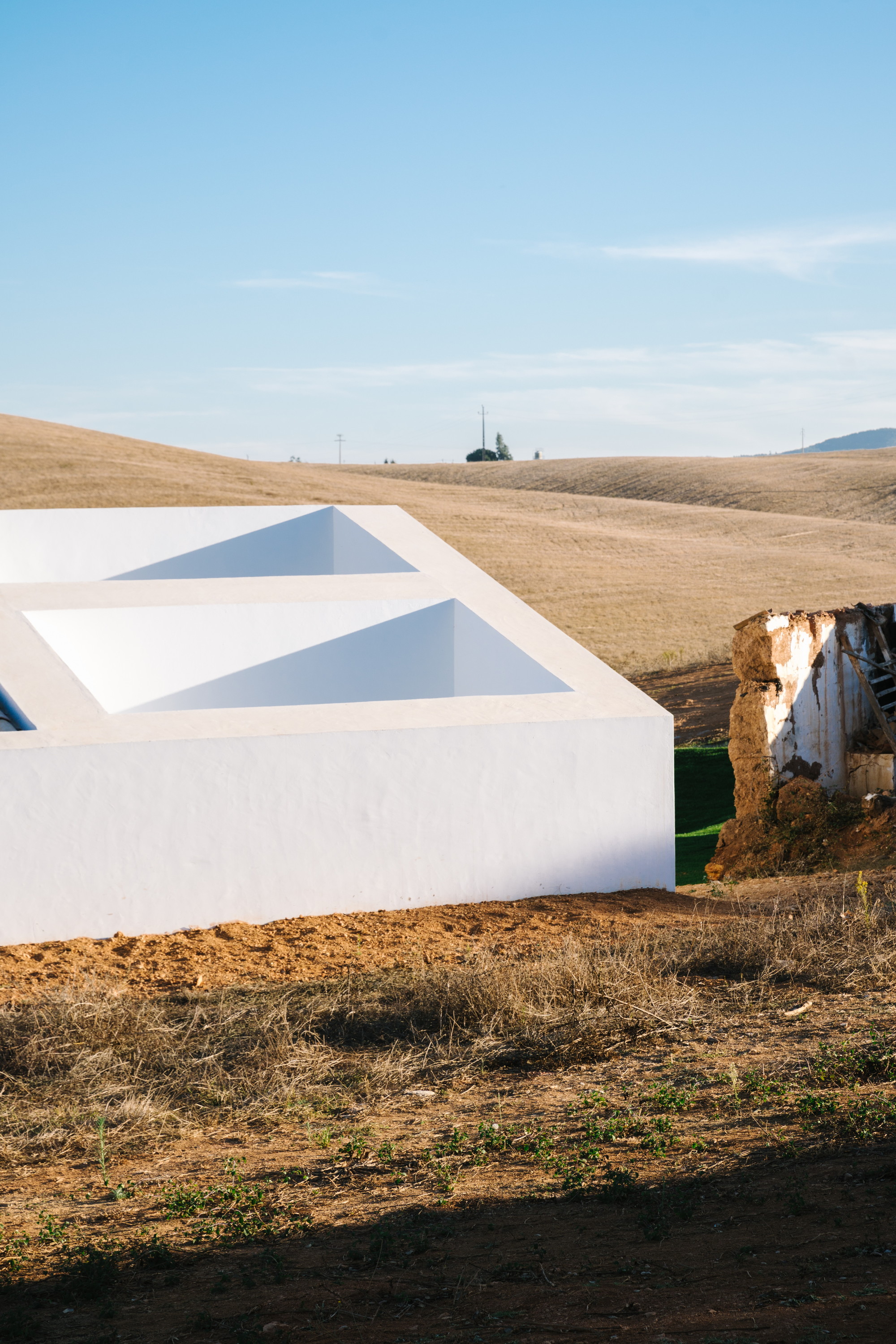 Cercal House by Atelier Data — A rendered white structure with pitched roof emerges from undulating agricultural terrain in the Alentejo landscape.