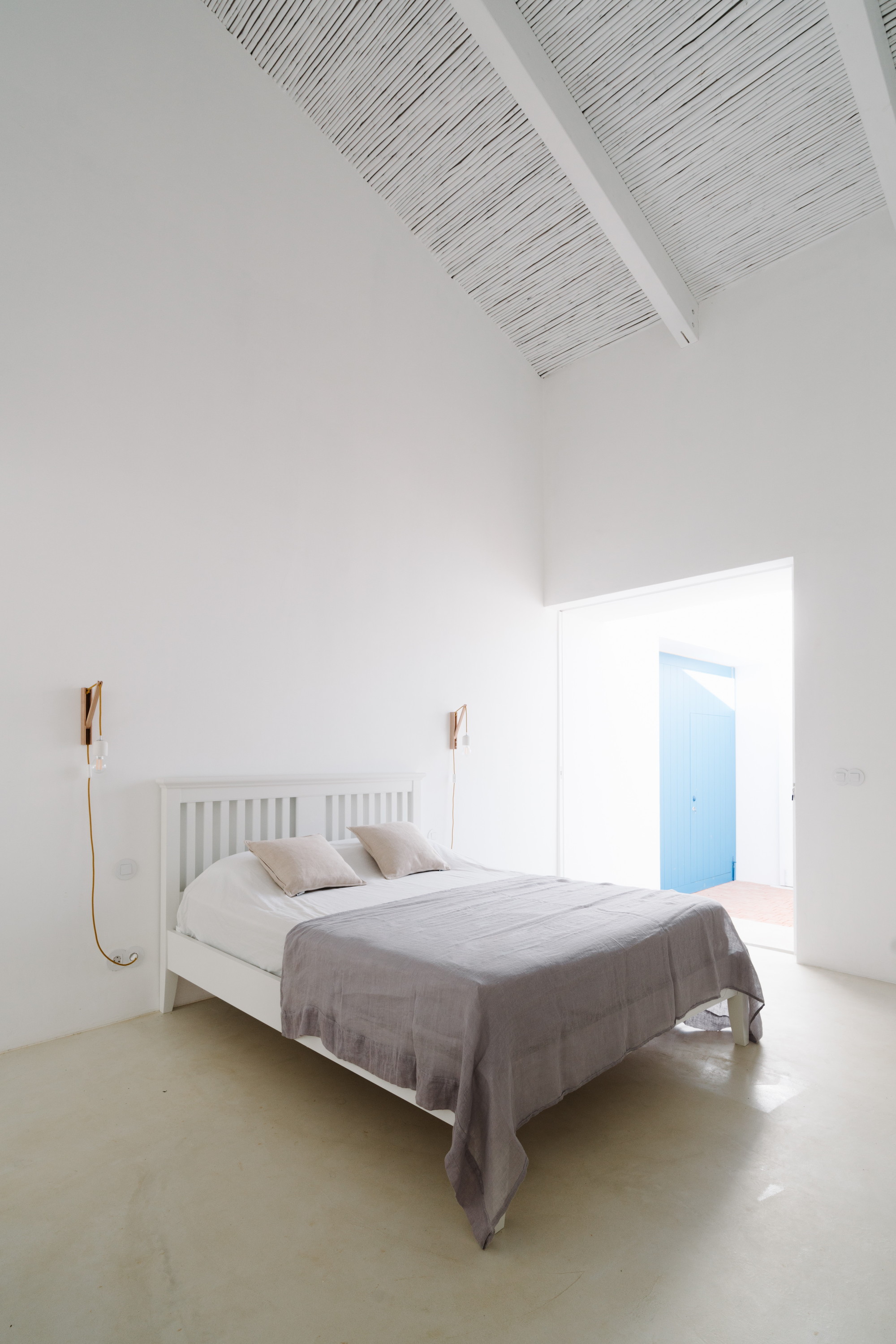 Cercal House by Atelier Data — A minimal bedroom interior flooded with natural light from a large glazed doorway revealing a bright blue door frame beyond.
