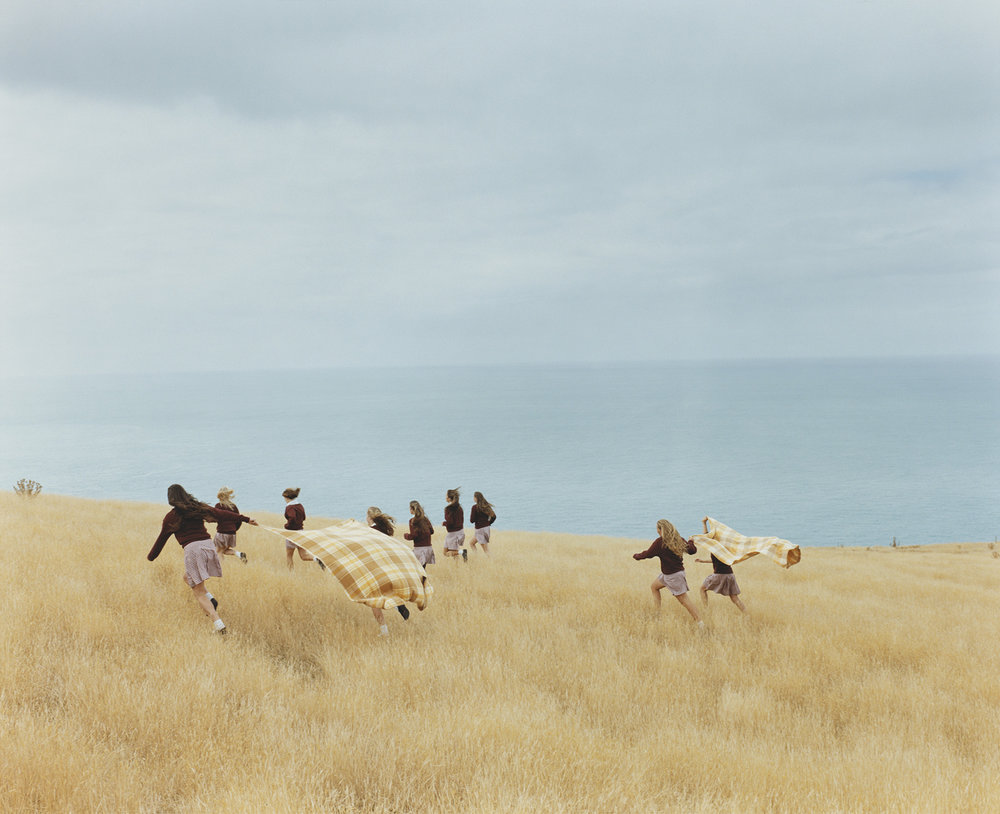 Assembly by Osamu Yokonami — Schoolgirls in dark uniforms and white pleated skirts traverse a dry grassland toward a calm turquoise sea.
