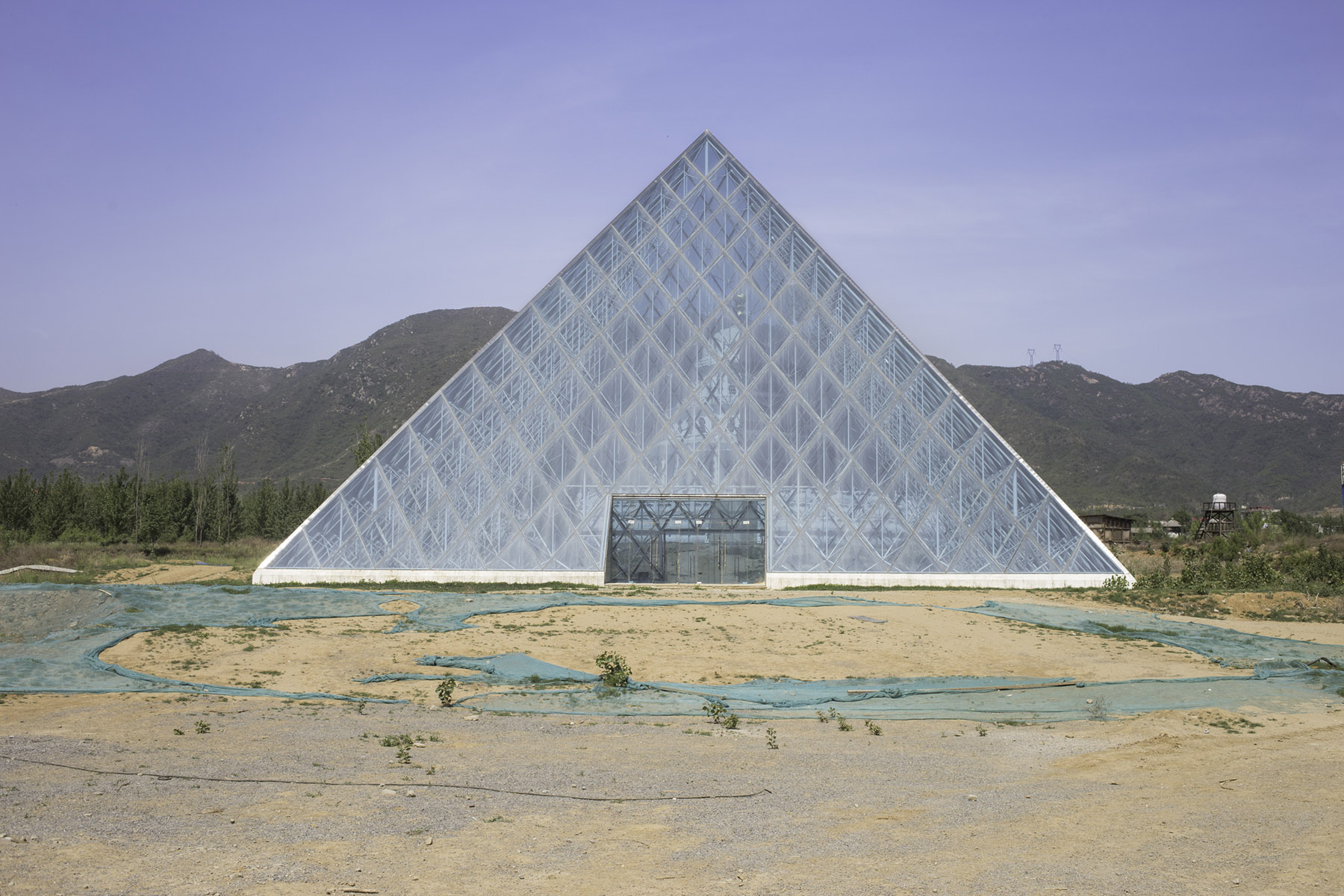 I am just a scenic spot by Pauilne Niks — A geometric glass pyramid structure dominates the arid landscape, its white framework and diamond-patterned glazing creating a volumetric form against...