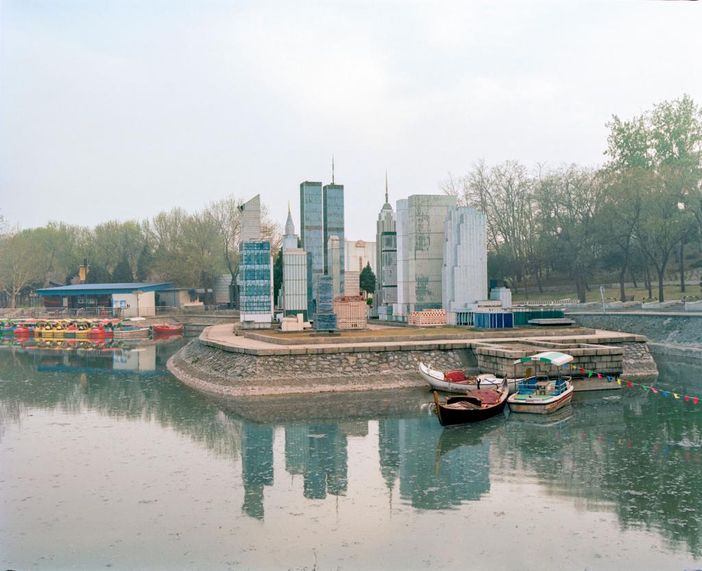 I am just a scenic spot by Pauilne Niks — A constructed miniature skyline occupies a peninsular island within a still waterway, featuring replica high-rise towers in concrete and glass rising behind...