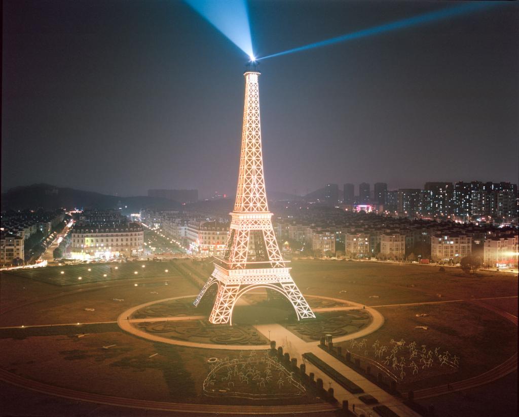 I am just a scenic spot by Pauilne Niks — A full-scale replica of the Eiffel Tower illuminated at night dominates a vast landscaped plaza in a Chinese city.