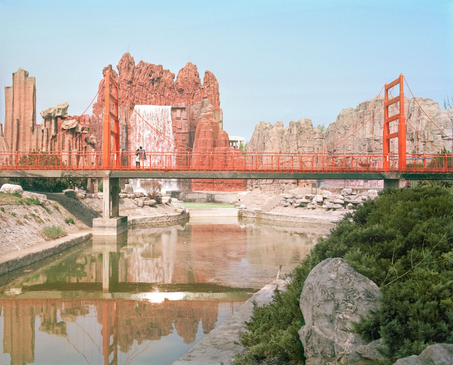 I am just a scenic spot by Pauilne Niks — A red-painted steel suspension bridge spans a silt-laden river, connecting reconstructed sandstone fortification walls with crenellated parapets.