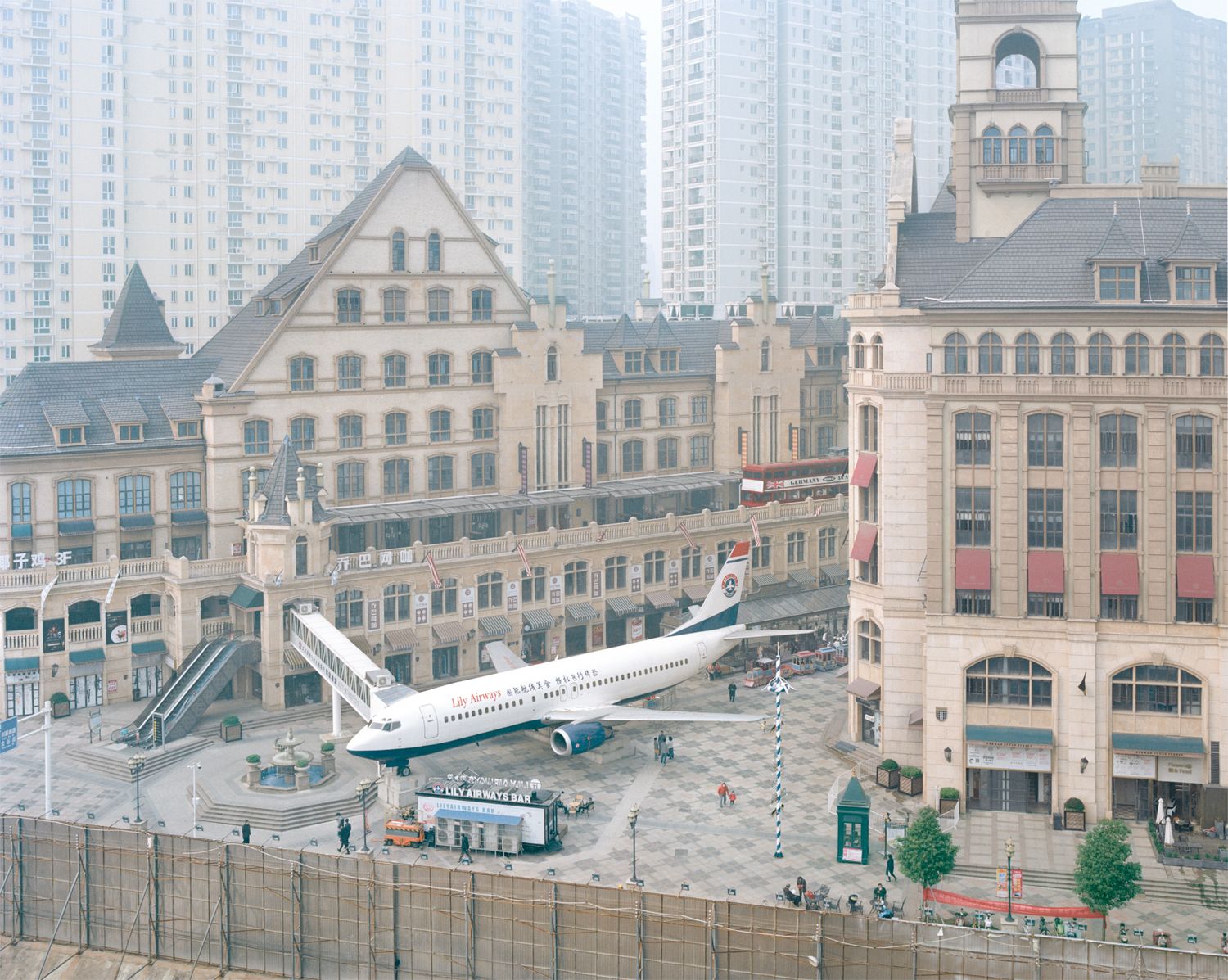I am just a scenic spot by Pauilne Niks — A Boeing 737 airliner is positioned in the central plaza of a constructed European townscape, surrounded by cream-rendered facades with slate-pitched roofs...