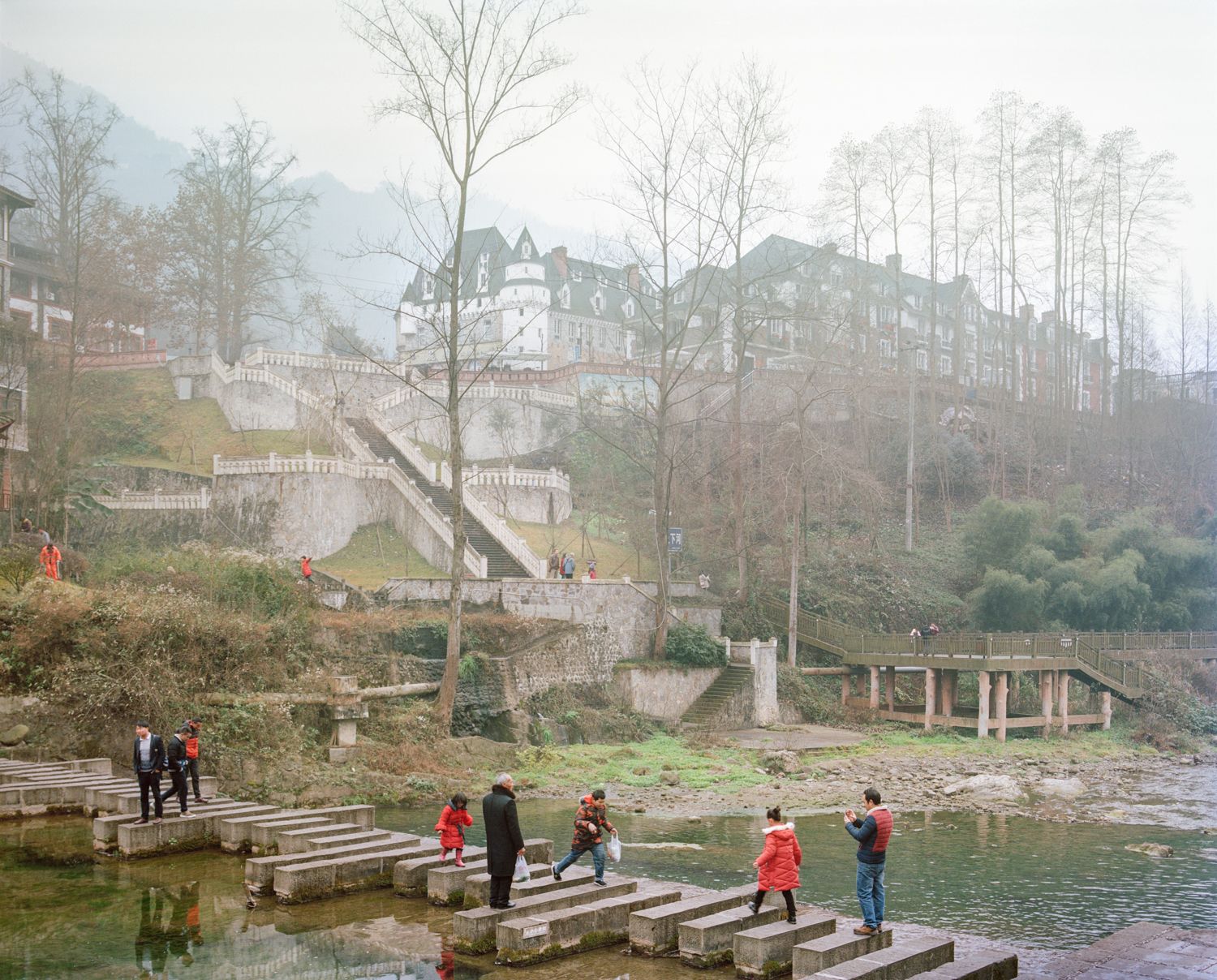 I am just a scenic spot by Pauilne Niks — Reconstructed stone fortification dominates the composition, its crenellated walls and stepped access points occupied by visitors in bright outerwear.