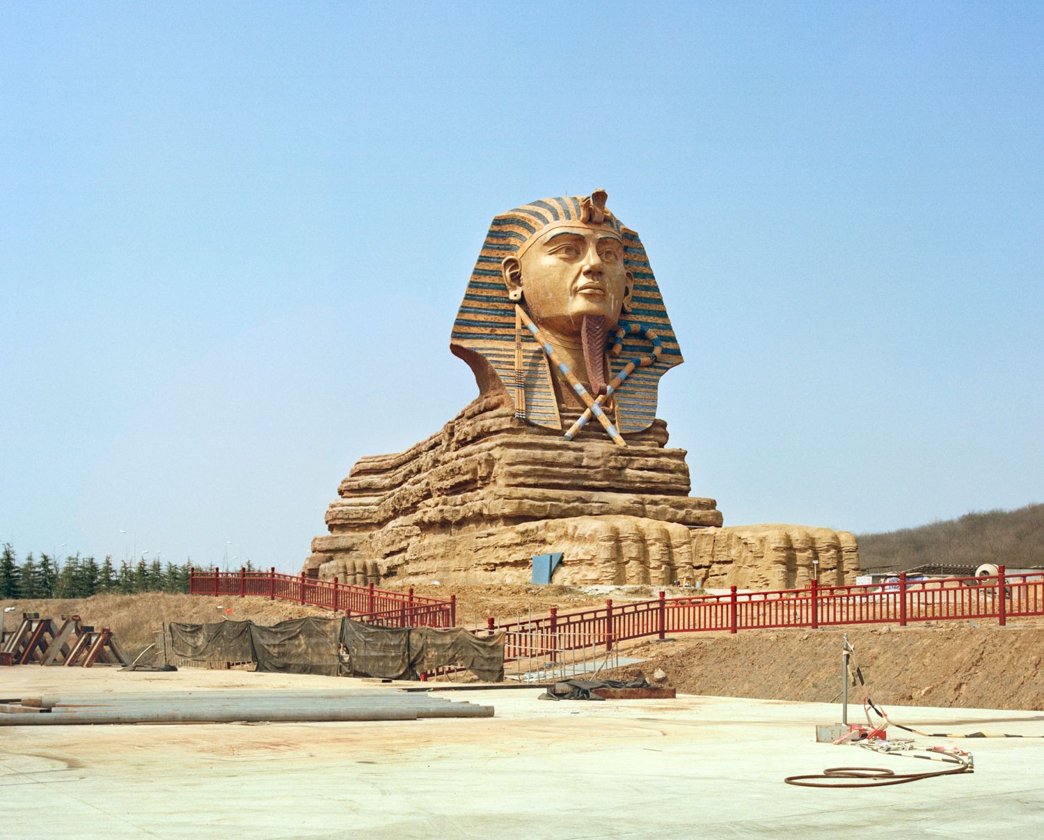 I am just a scenic spot by Pauilne Niks — A colossal pharaonic head sculpture dominates an active construction site in rural China, its gilded stone visage gazing upward against clear sky.