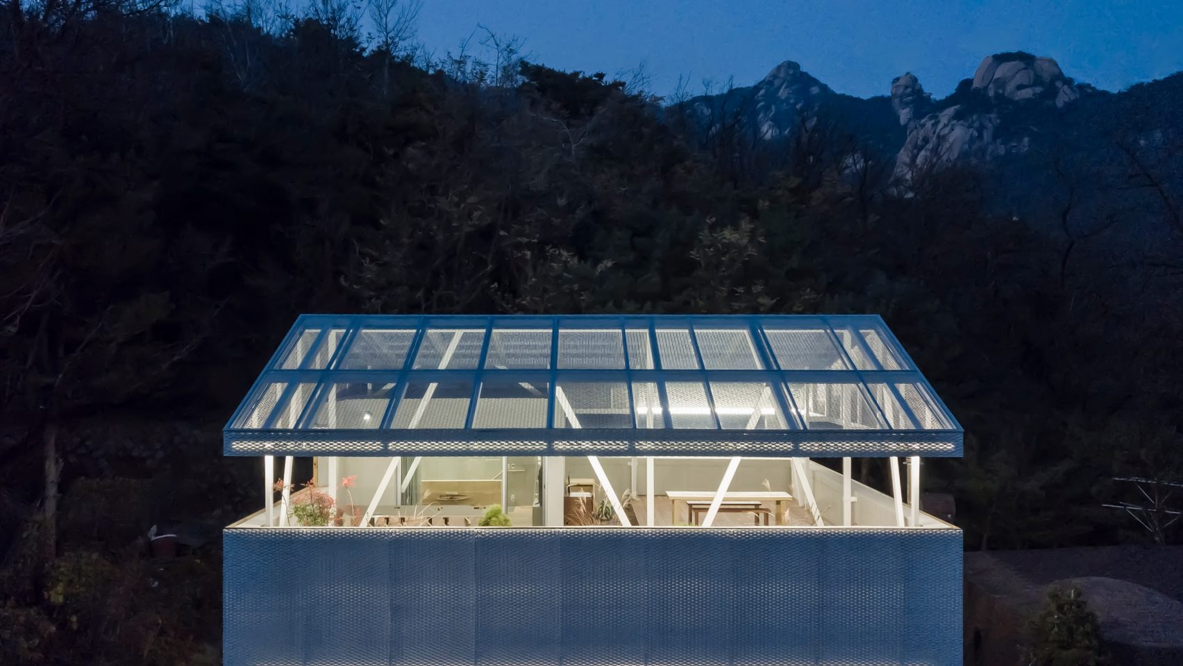 The Shaded Rooftop House by BCHO Architects — Twilight photography captures the Shaded Rooftop House as a luminous pavilion against darkened mountainous terrain.