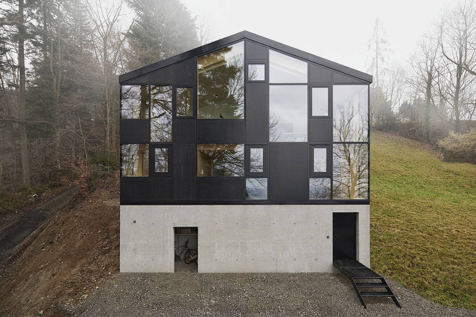 House Hohlen by Jochen Specht — House Hohlen presents a two-part volumetric composition: a recessed concrete plinth supporting an elevated timber-framed upper volume clad in black panels...
