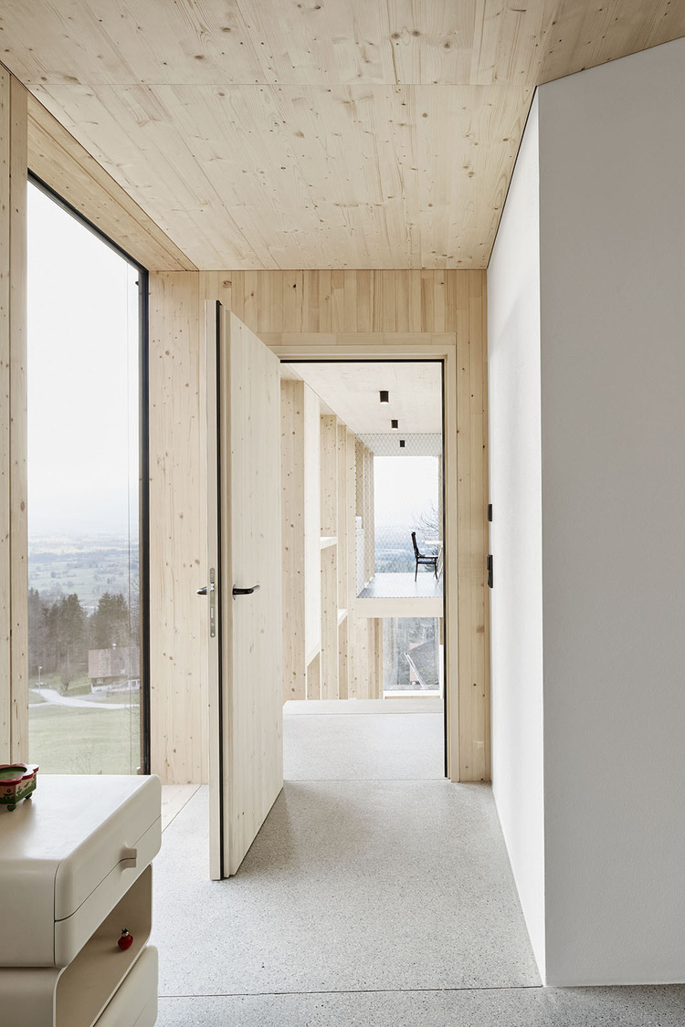 House Hohlen by Jochen Specht — A corridor passage lined with timber board-and-batten walls and ceiling runs perpendicular to a large black-framed glass panel overlooking the Austrian...