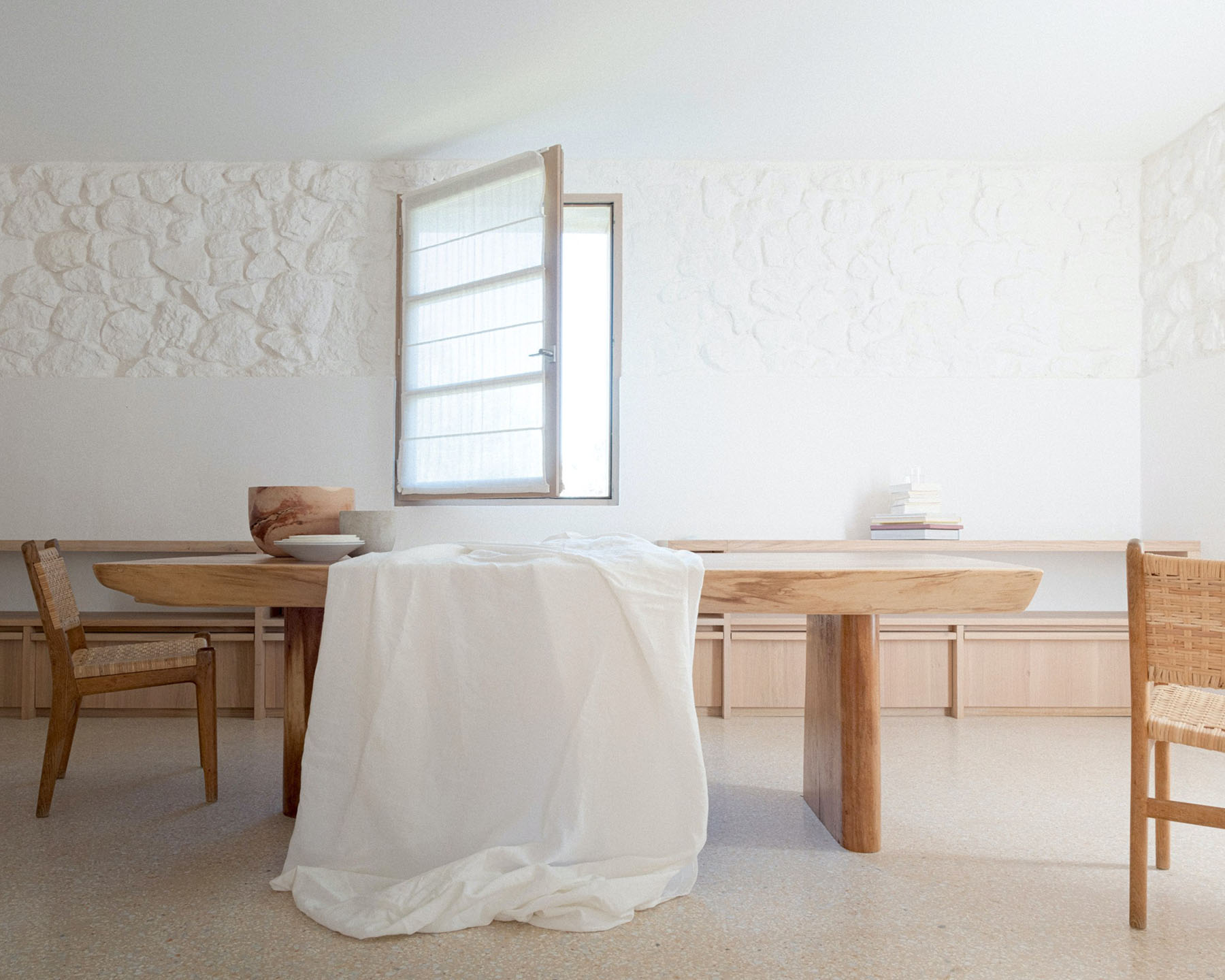 MA House by Studio XM — Interior view of a minimalist dining space featuring a substantial wooden table with cylindrical support, draped in cream linen.