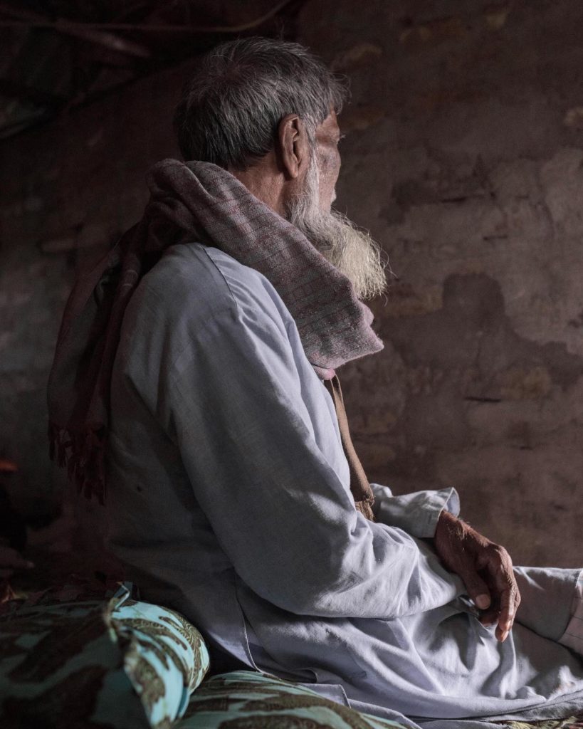 Ebbing away of Identity with the Tides by Sushavan Nandy — A seated elderly man with gray hair and long beard faces away from the camera in profile, positioned against a weathered brown plaster wall.