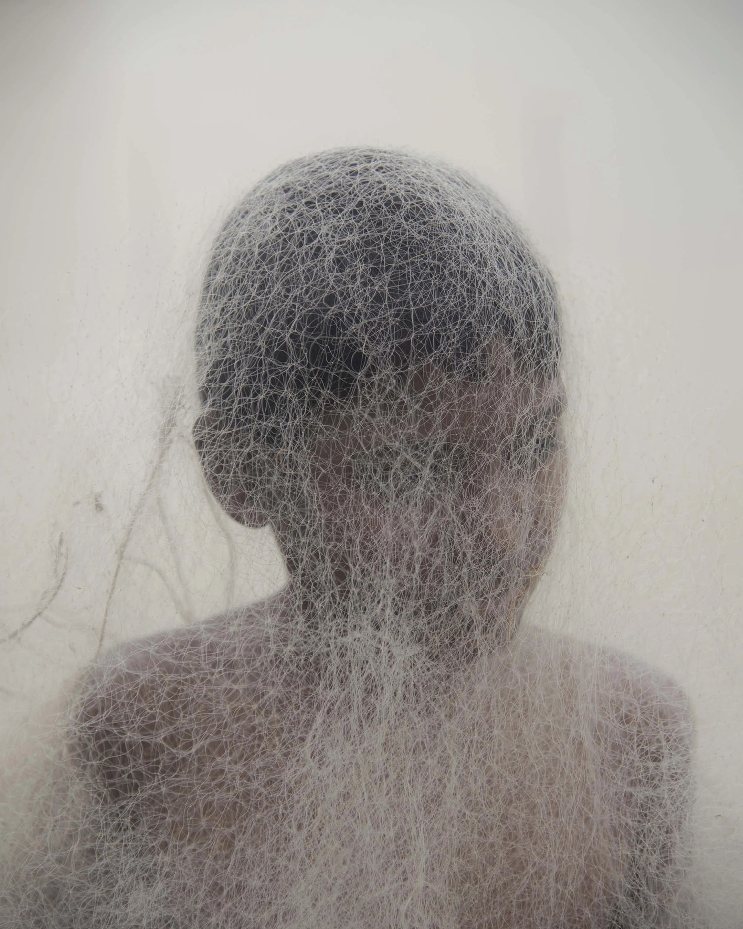 Ebbing away of Identity with the Tides by Sushavan Nandy — A translucent sculptural form constructed from fine mesh or thread woven into a human head and shoulder silhouette.