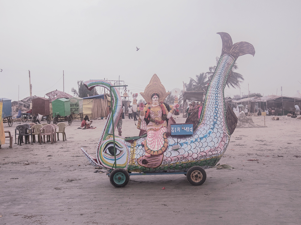 Ebbing away of Identity with the Tides by Sushavan Nandy — A sculptural wooden boat-shaped vehicle dominates the sandy plaza of what appears to be a coastal settlement in the Sundarbans.