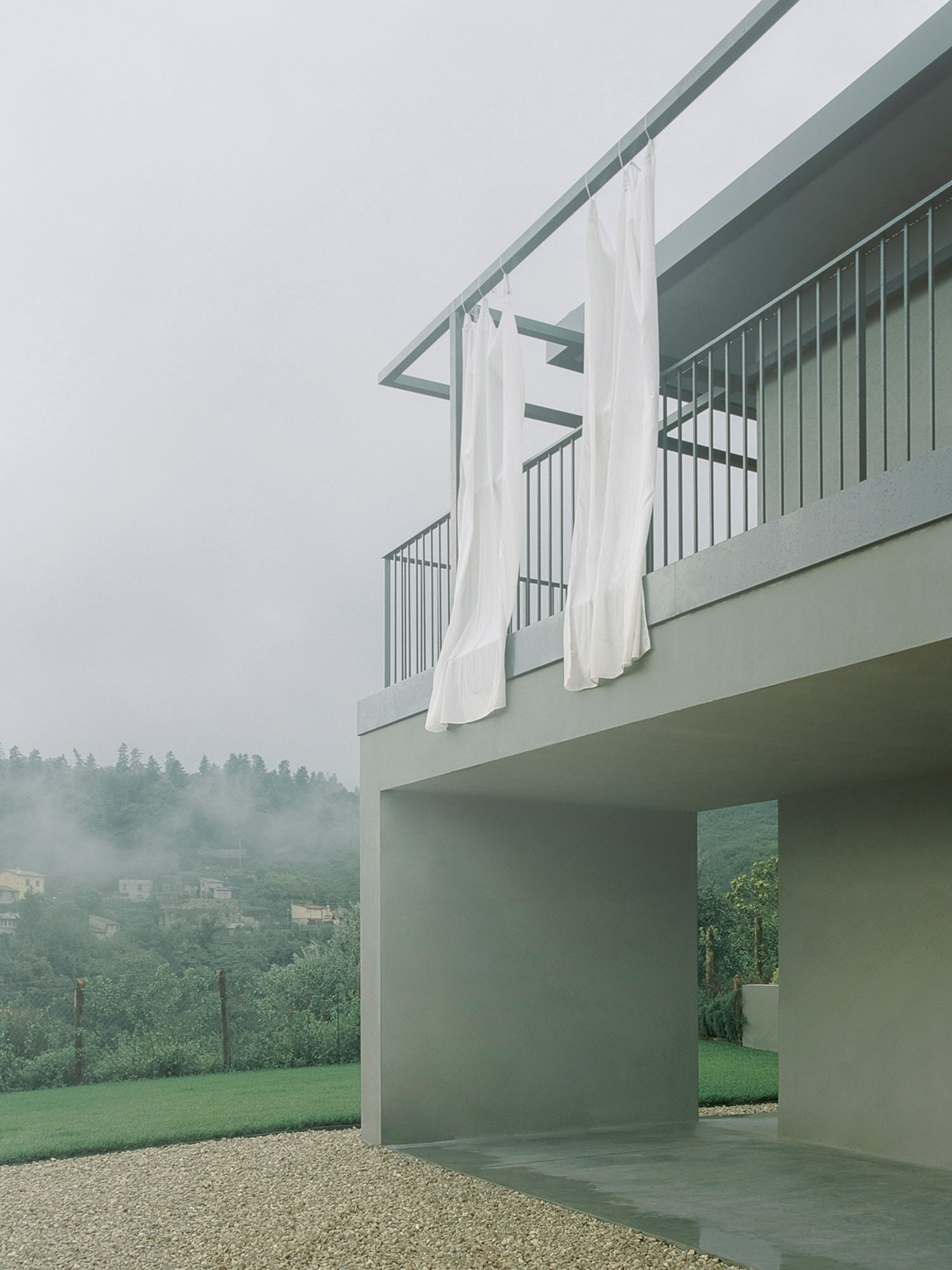 Elena by Deltastudio — A rendered concrete structure in sage-green occupies the lower volume, its clean horizontal planes interrupted by a cantilevered upper deck framed by...