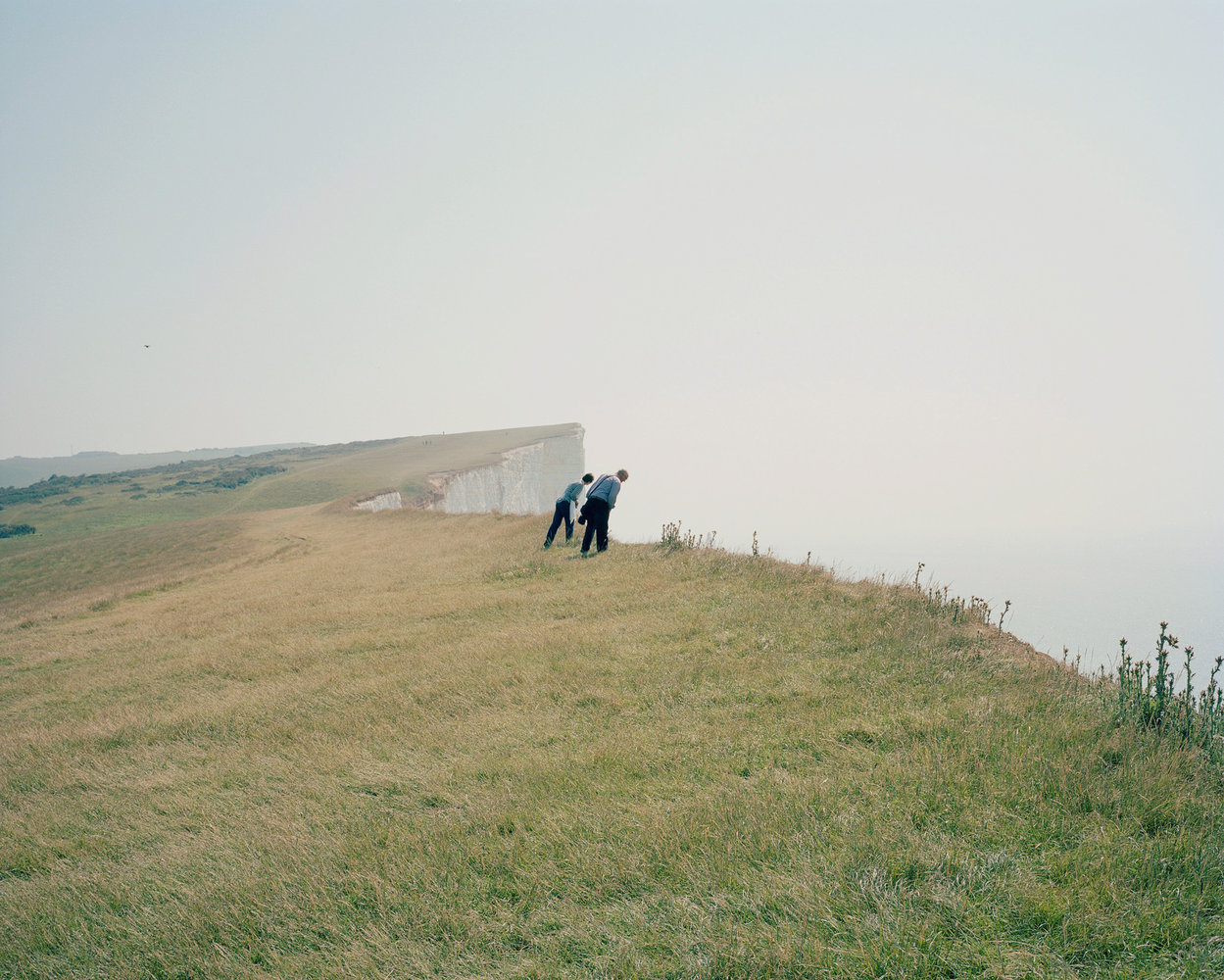 Selected works by Toby Coulson — Two figures in dark clothing traverse a sloping grassland toward a dramatic white chalk cliff face receding into the distance.