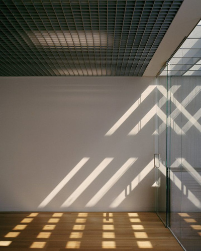 Yoga Studio by Richard Stampton — Gridded translucent ceiling panels cast a uniform pattern of light across the white wall and timber floor, creating a geometric shadow composition.