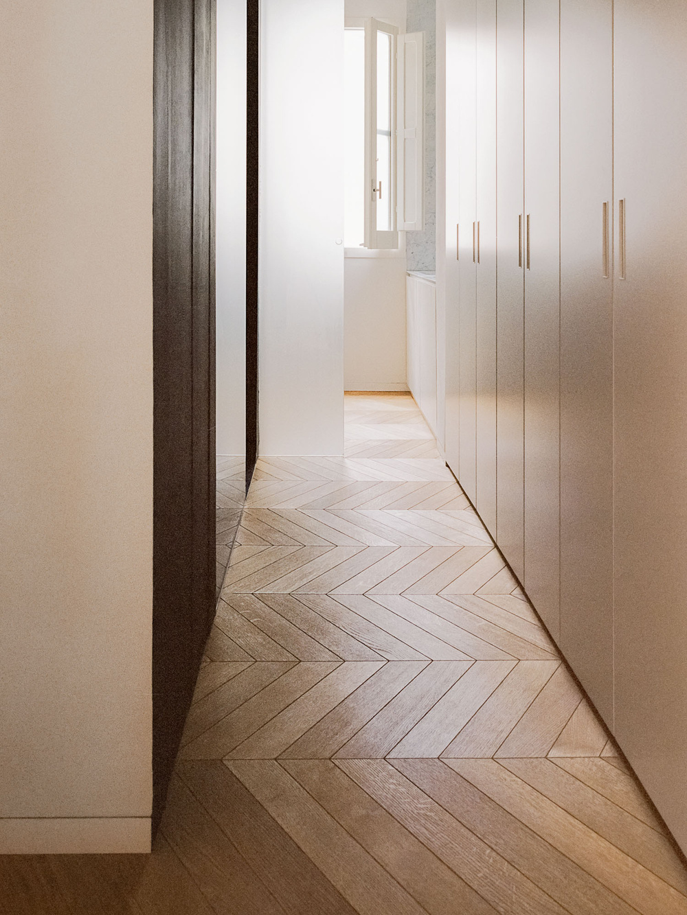 House for a Couple by SET Architects — A corridor passage with herringbone oak flooring in mixed light and dark tones establishing directional rhythm toward a distant threshold.