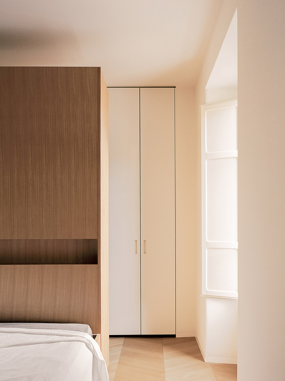 House for a Couple by SET Architects — A narrow bedroom passage frames a tall cream-lacquered wardrobe with brass handles, its two-door configuration rising to meet a sloped ceiling.