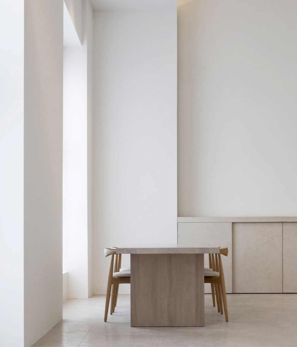 Alkema Studio by Jen Alkema Architect — A dining ensemble anchors the center of a minimalist interior, featuring a monolithic timber table with faceted sides positioned between two substantial...