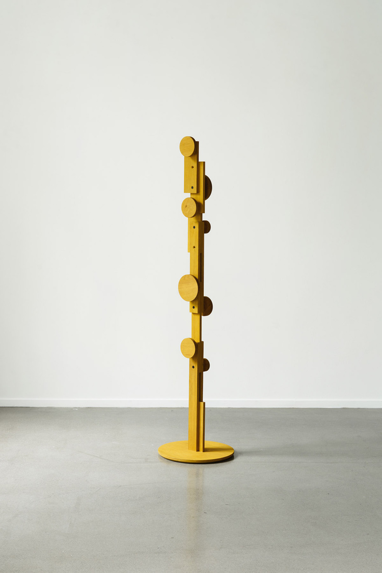 Unika Auction by Bjørn van den Berg and Falke Svatun — A tall wooden coat stand in yellow ochre finish, featuring a vertical rectangular post mounted on a circular base.
