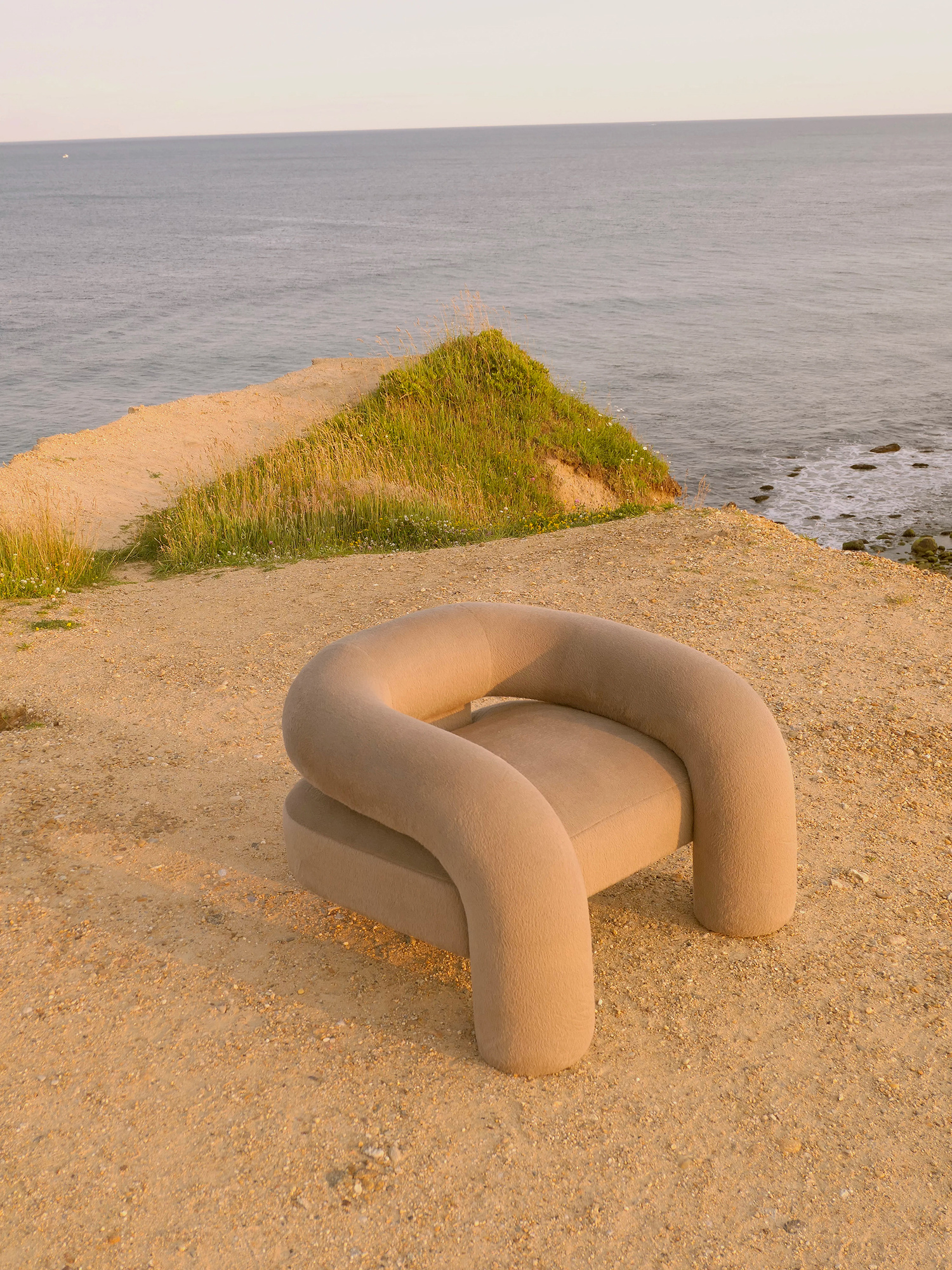 Kosa Collection by Ian Felton — A sculptural chair with continuous tubular forms sits on a sandy beach overlooking calm water.